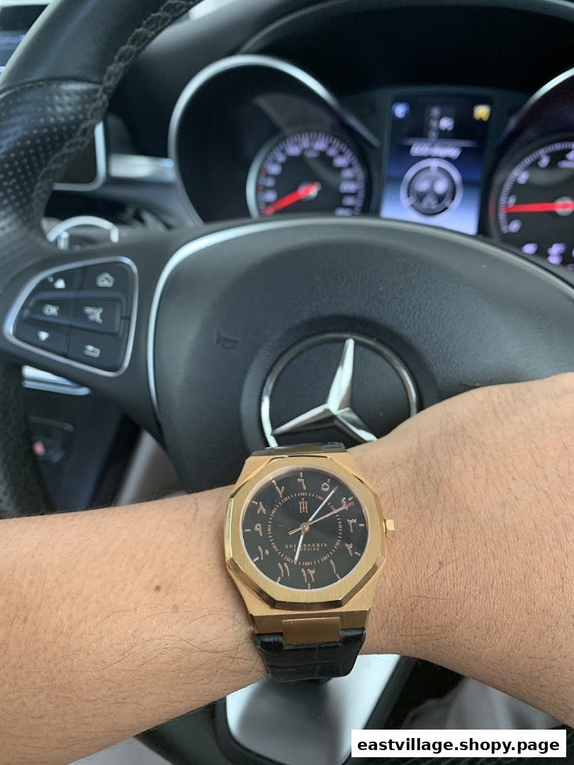 A person's arm wearing a gold watch with Arabic numerals, inside a car with a Mercedes-Benz steering wheel.