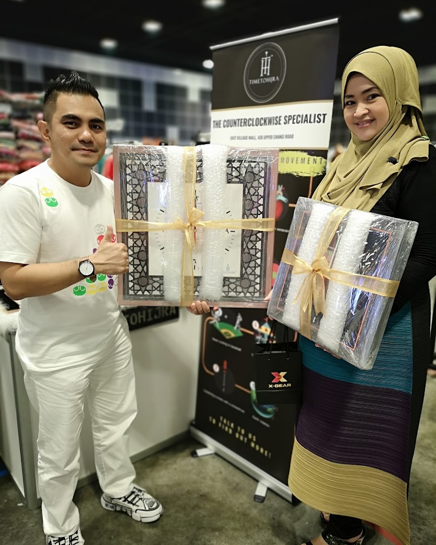 Two individuals proudly display packaged counterclockwise clocks from 'Time To Hijra' at their booth.