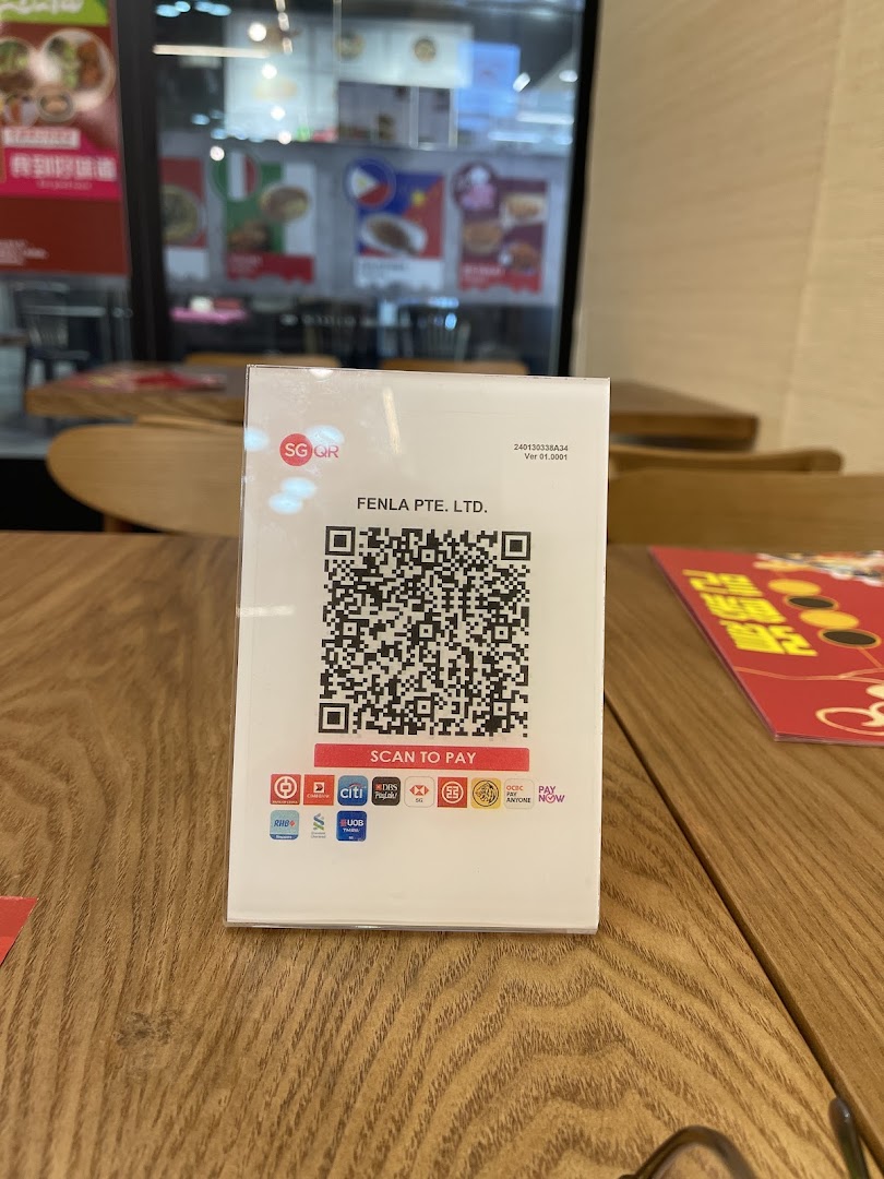 A close-up of an SG QR payment stand on a wooden table inside a restaurant, with food posters in the background.