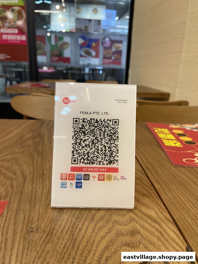A close-up of an SG QR payment stand on a wooden table inside a restaurant, with food posters in the background.