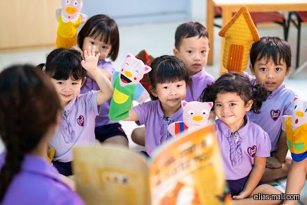 Children with puppets listen to a teacher reading a book in a classroom.