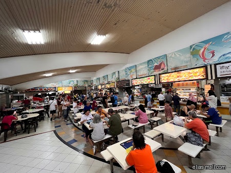 The image shows a busy food court with many people and food stalls.
