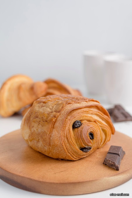 A pain au chocolat pastry sits on a wooden board with chocolate pieces.