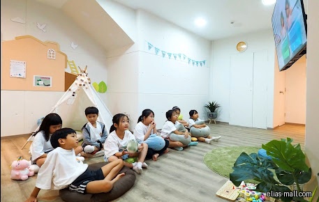 Children sit watching a screen in a bright, decorated classroom setting.