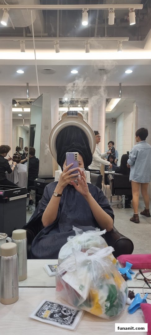 A customer takes a selfie while receiving a hair treatment with a steamer at a salon.