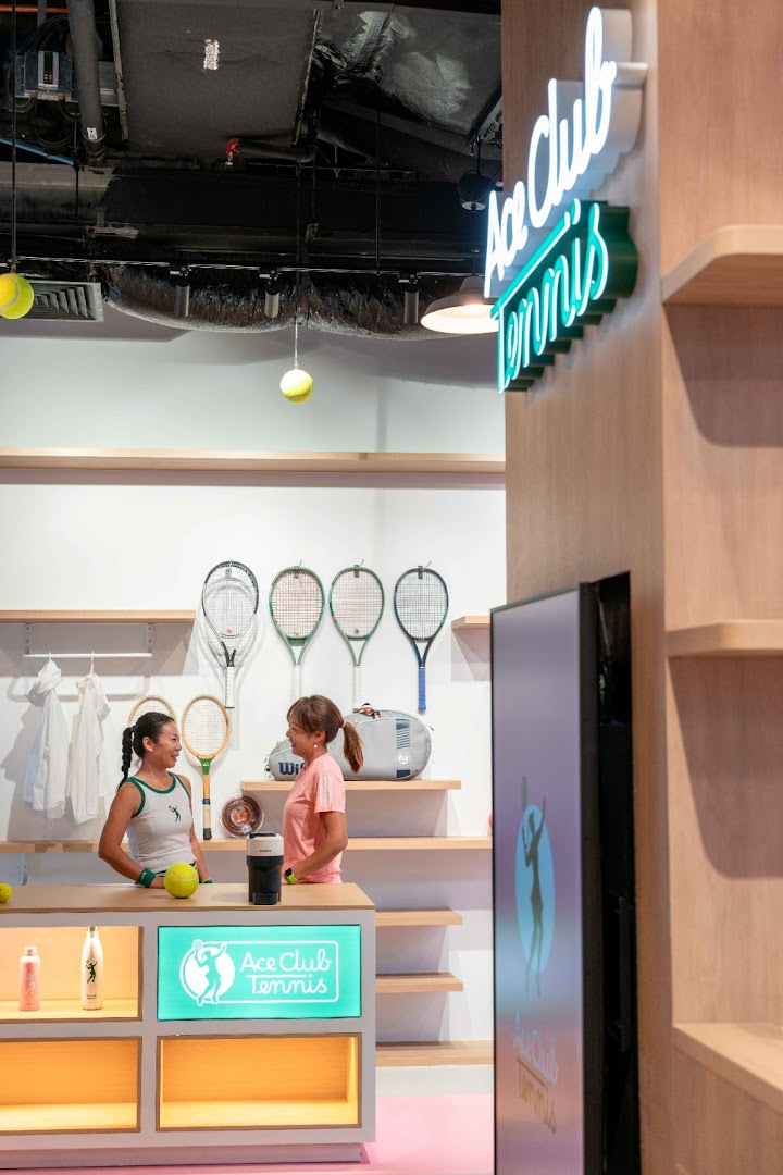 Two women converse inside Ace Club Tennis, surrounded by tennis rackets and merchandise.