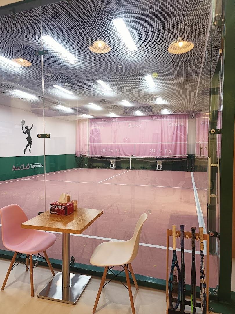 An indoor tennis court with a pink surface and a small seating area, viewed through a glass partition.