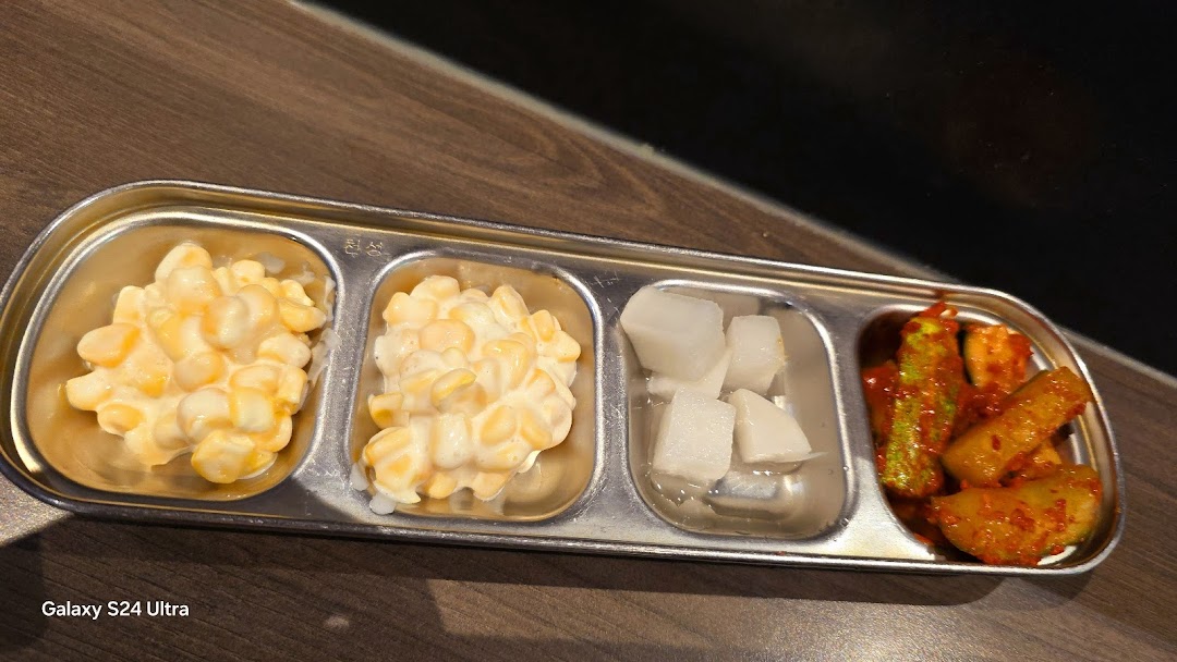 A metal tray with four compartments holding Korean banchan, including corn salad, pickled radish, and spicy kimchi.