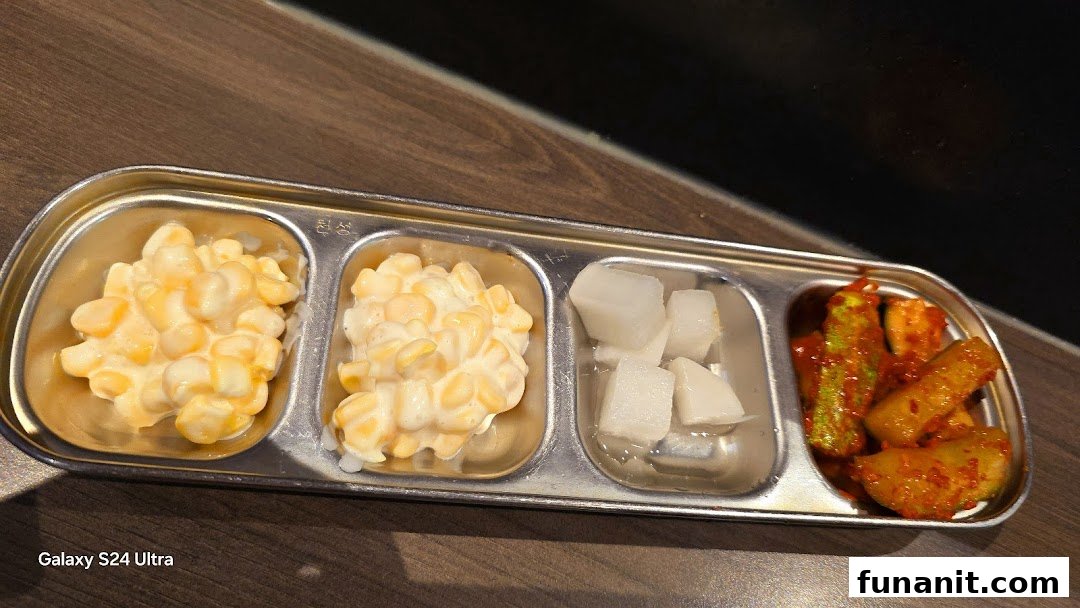 A metal tray with four compartments holding Korean banchan, including corn salad, pickled radish, and spicy kimchi.