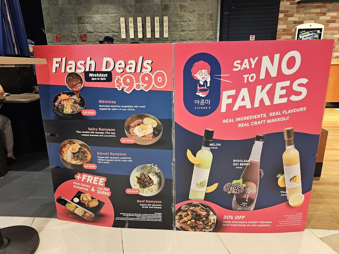 A promotional standee for Ajumma's, showcasing Korean food deals and authentic makgeolli drinks.