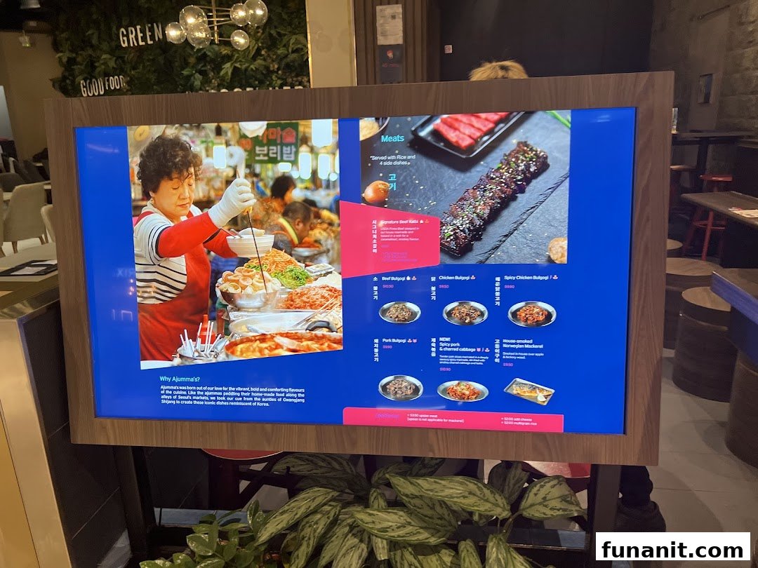 A digital menu display for Ajummas, showcasing Korean dishes and a market scene.