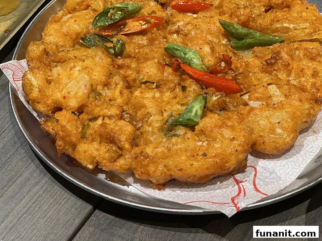 A close-up of a golden-fried dish garnished with red and green chili peppers and herbs on a metal plate.