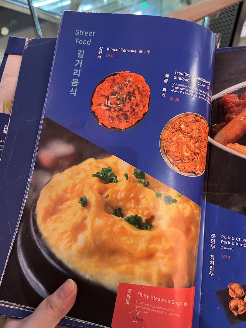A close-up view of a Korean restaurant menu, featuring street food items like pancakes and steamed eggs.