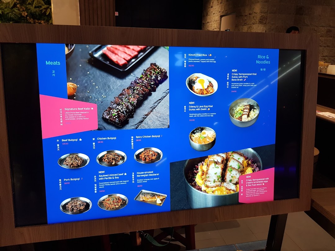 A digital menu board at Ajummas displaying various Korean dishes and their prices.