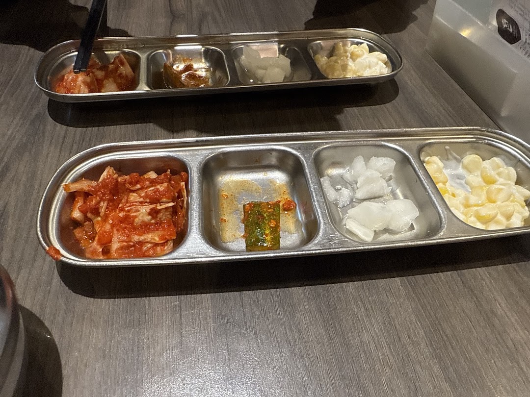 Two metal trays filled with various Korean banchan, including kimchi and corn salad, on a wooden table.