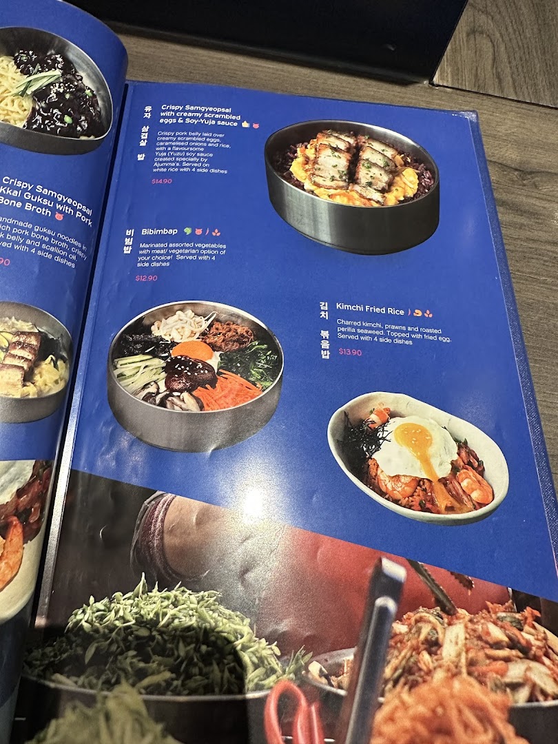A vibrant menu page from Ajummas, showcasing various Korean dishes with descriptions and prices.