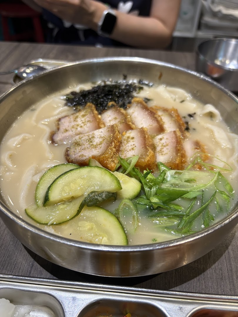 A close-up of a delicious bowl of Korean noodles with crispy pork belly, vegetables, and seaweed.