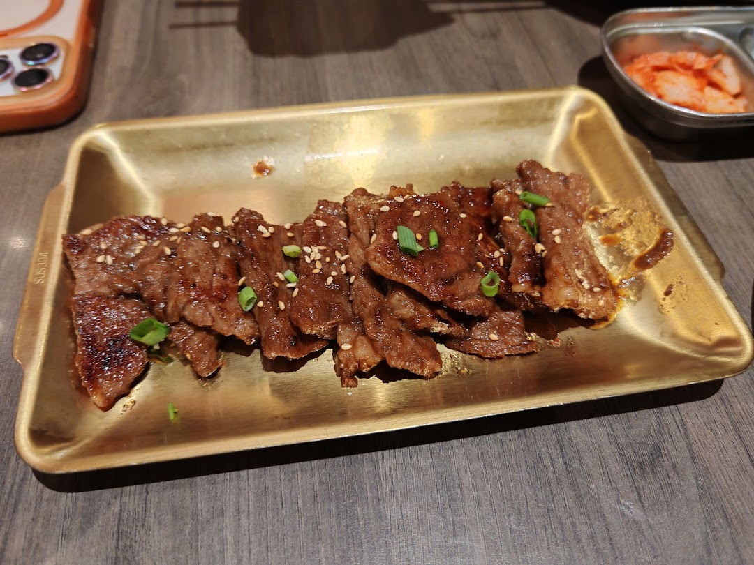 A golden tray holds slices of grilled marinated meat, garnished with sesame seeds and green onions, alongside a dish of kimchi.