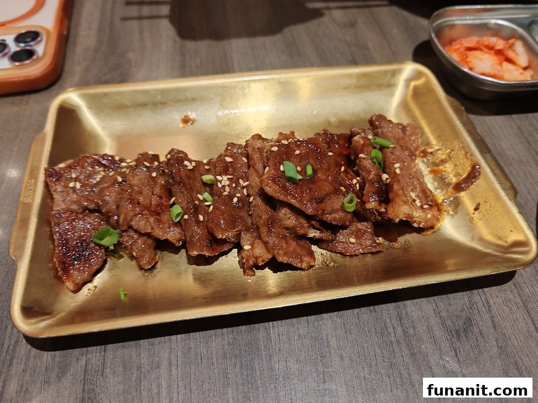 A golden tray holds slices of grilled marinated meat, garnished with sesame seeds and green onions, alongside a dish of kimchi.