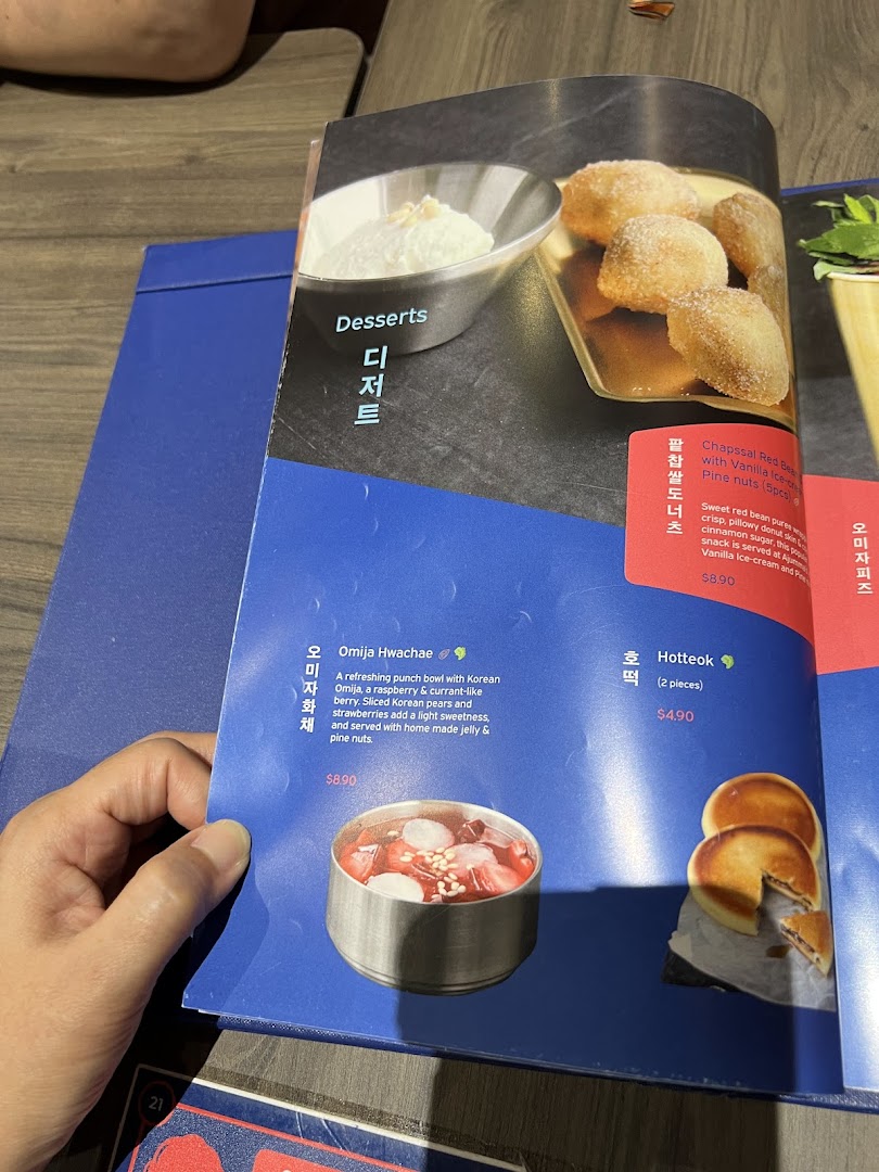 A hand holds open a menu page displaying Korean desserts with descriptions and prices.