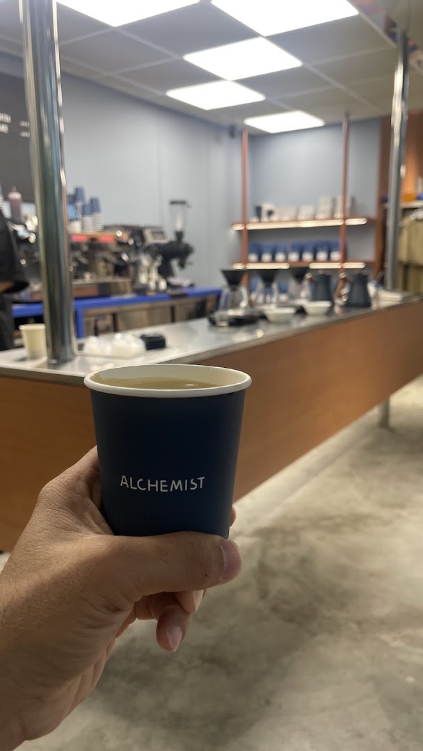A hand holds an Alchemist branded coffee cup inside a modern coffee shop with brewing equipment visible.