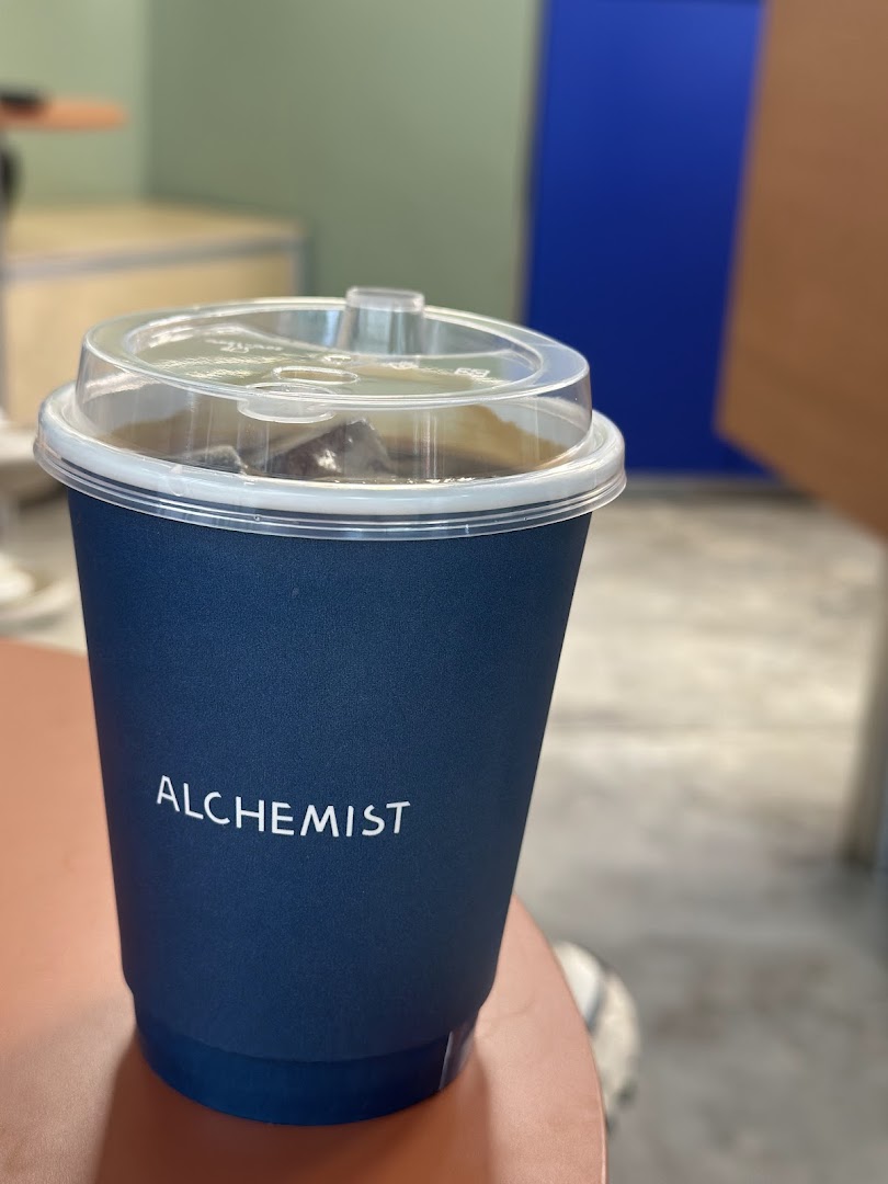 A dark blue Alchemist coffee cup with a clear lid, containing an iced beverage.