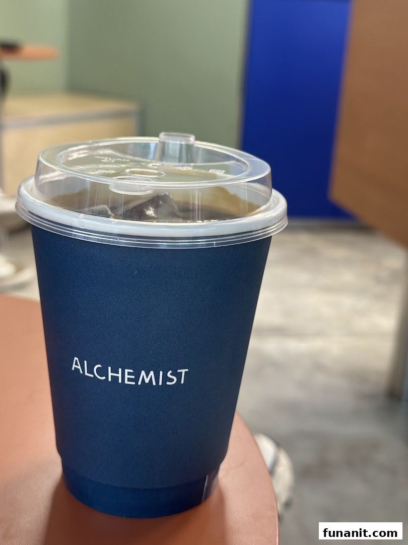 A dark blue Alchemist coffee cup with a clear lid, containing an iced beverage.