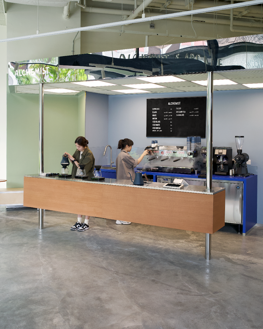 Two baristas preparing coffee at a modern, minimalist coffee bar with a menu board.