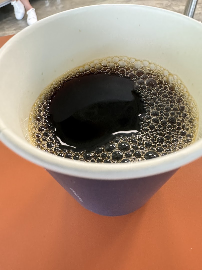 A close-up view of a dark beverage with a frothy top in a paper cup.