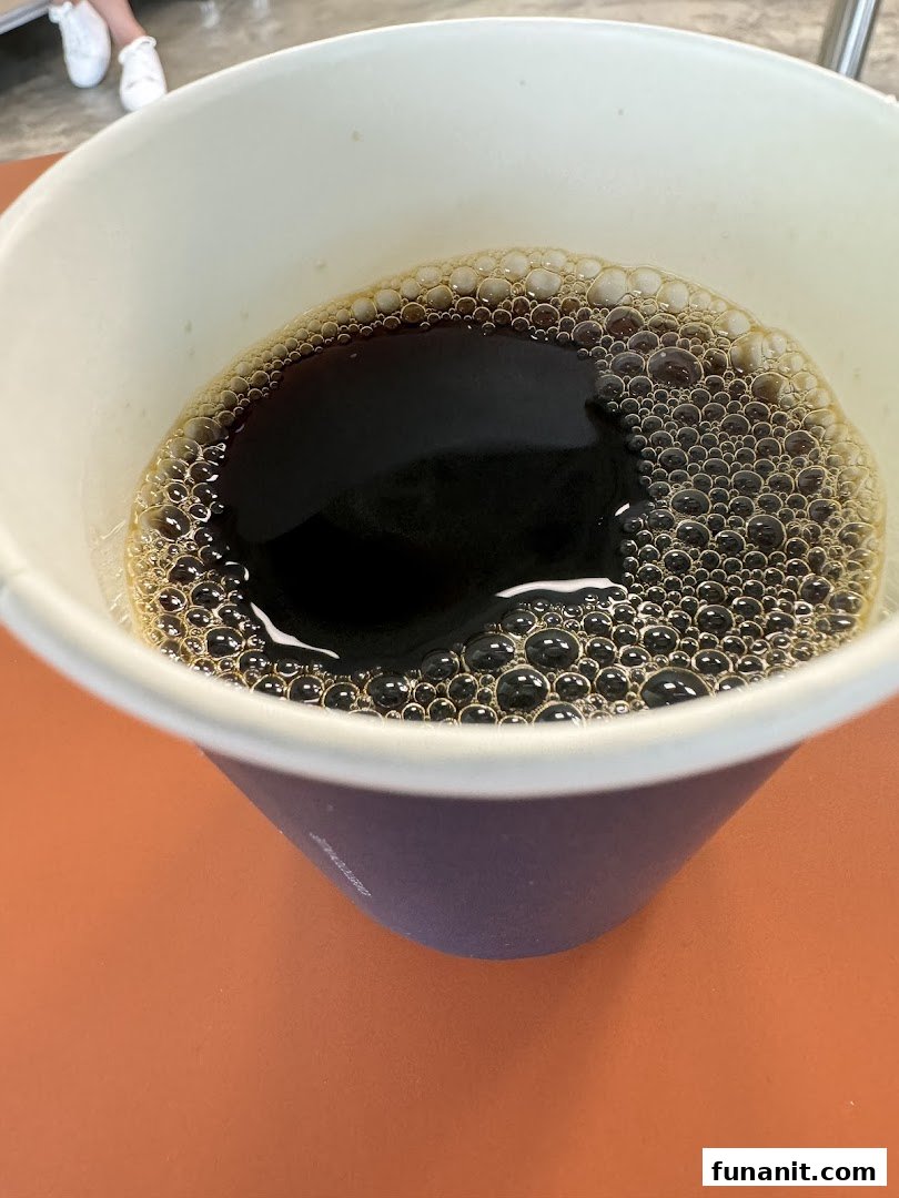 A close-up view of a dark beverage with a frothy top in a paper cup.