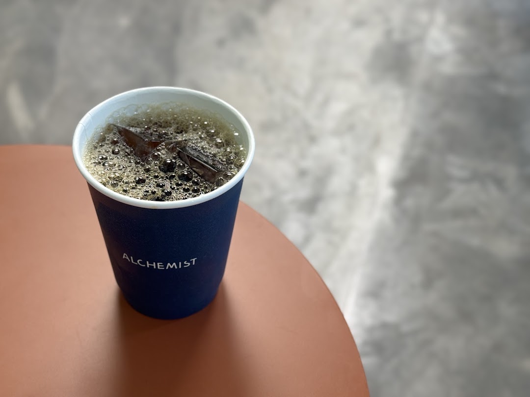 A dark blue cup of iced coffee with the 'Alchemist' logo on a reddish-brown table.