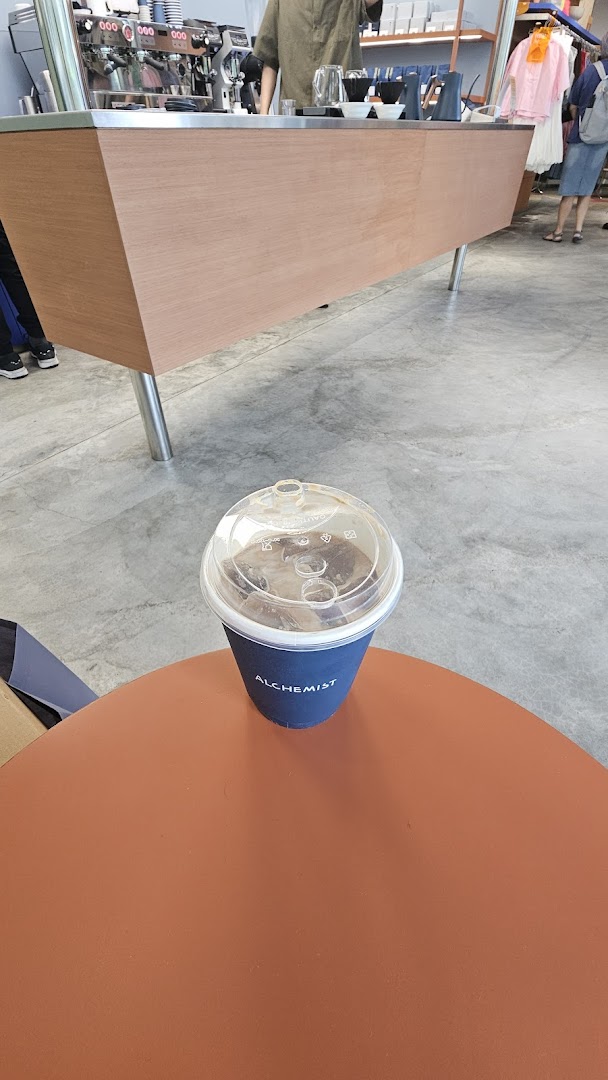 An iced coffee from Alchemist Funan sits on a table, with the coffee bar and staff in the background.