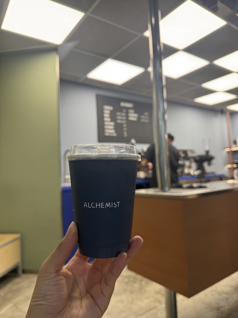 A hand holds an Alchemist branded coffee cup inside the shop, with the counter and menu board in the background.