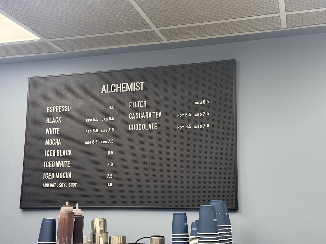 A black menu board displaying coffee and specialty drink options with prices at Alchemist.