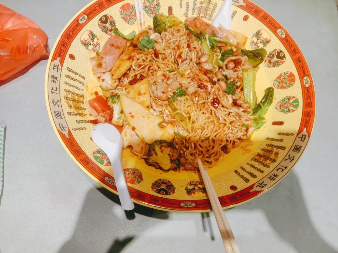 A large, colorful bowl of spicy noodles with various ingredients, served with chopsticks and a spoon.