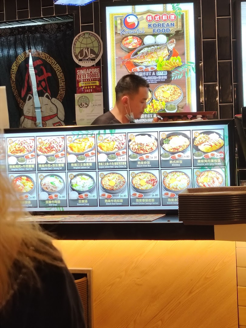 A staff member stands behind a counter displaying various Korean food menu items and prices.