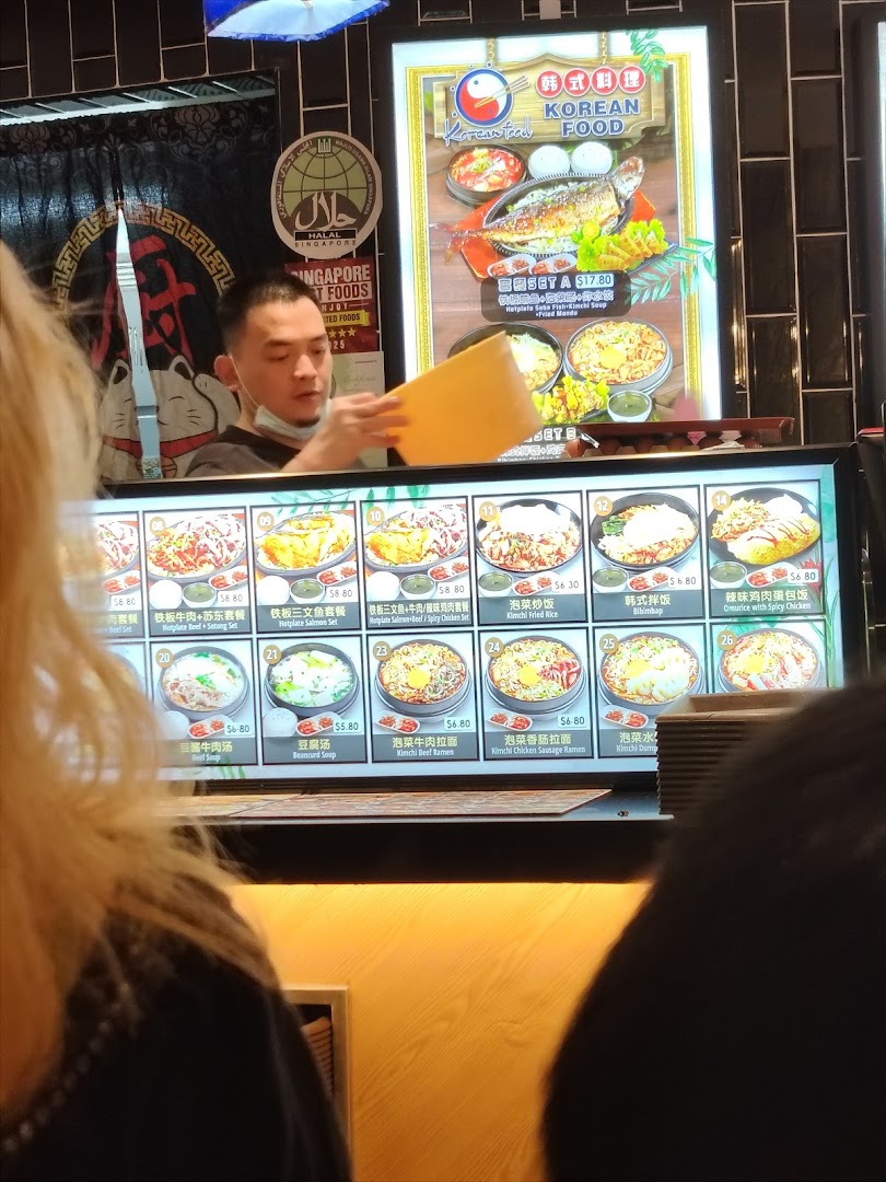 A staff member stands behind a counter with a digital menu displaying various Korean food items and prices.