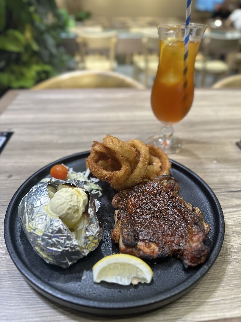 A grilled chicken steak with onion rings, baked potato, and a drink on a black plate at a restaurant.