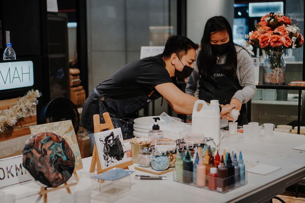 Two individuals in aprons and masks are engaged in a hands-on art activity at a table filled with supplies.