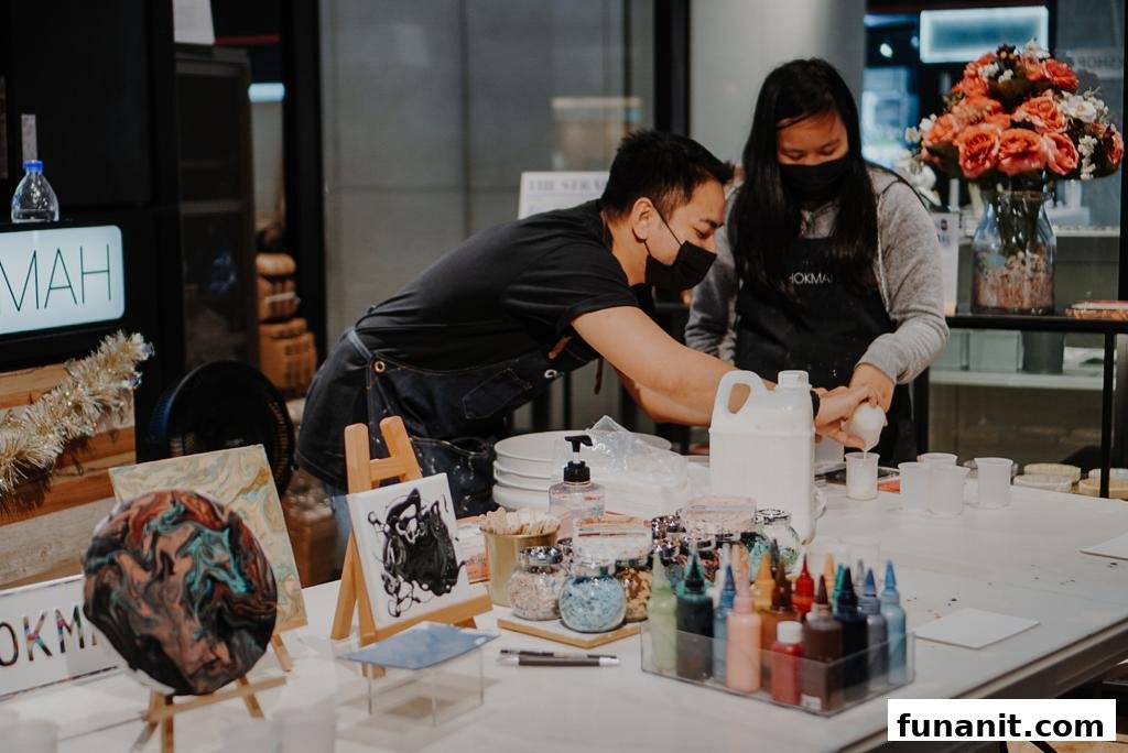 Two individuals in aprons and masks are engaged in a hands-on art activity at a table filled with supplies.
