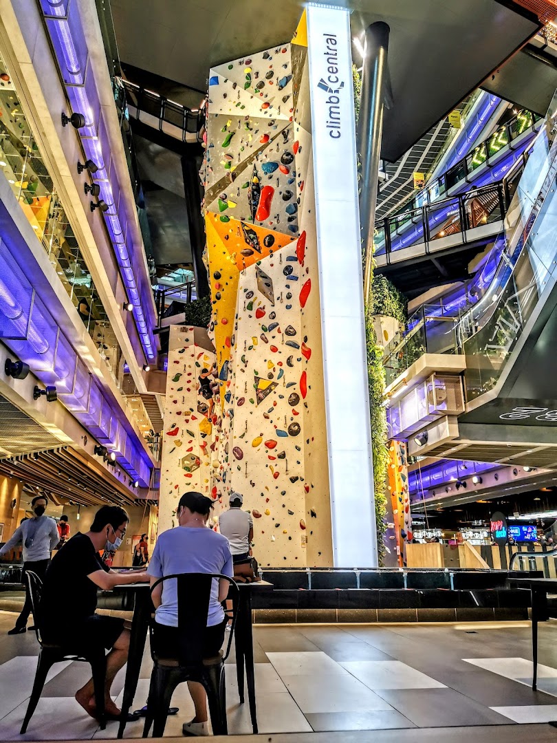 An indoor climbing gym with a tall, colorful climbing wall and people relaxing nearby.