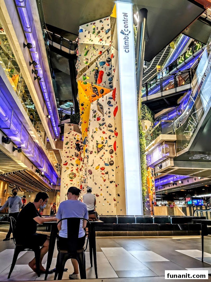 An indoor climbing gym with a tall, colorful climbing wall and people relaxing nearby.