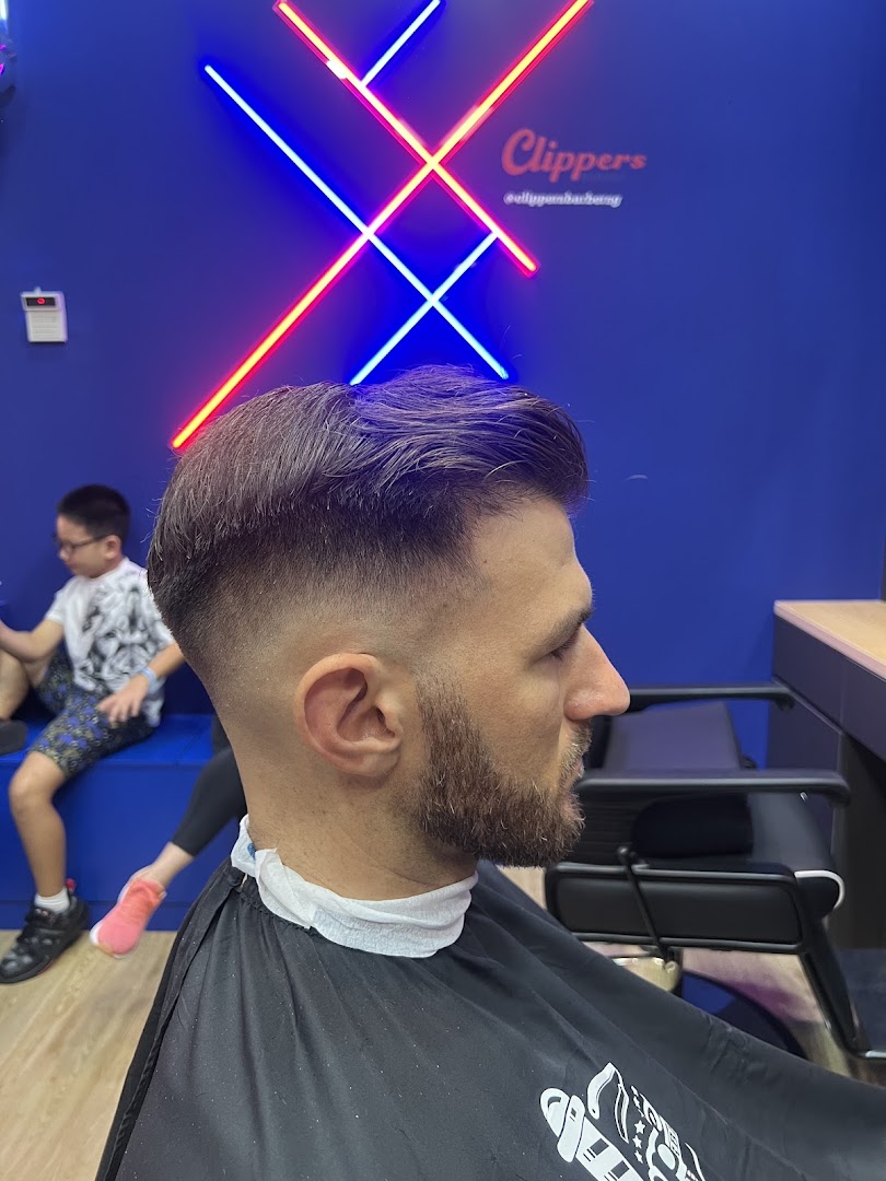 A man receiving a fresh haircut and beard trim inside Clippers Barber, with neon signage in the background.