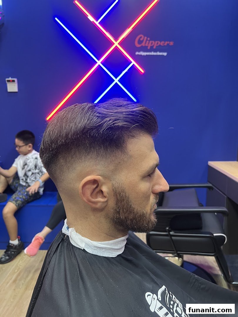 A man receiving a fresh haircut and beard trim inside Clippers Barber, with neon signage in the background.