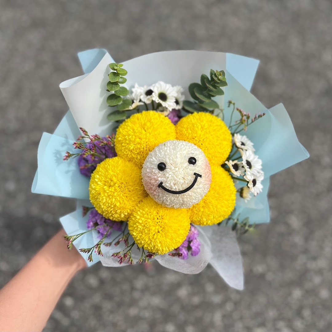 A hand holds a vibrant flower bouquet featuring a smiley face bloom surrounded by yellow flowers.