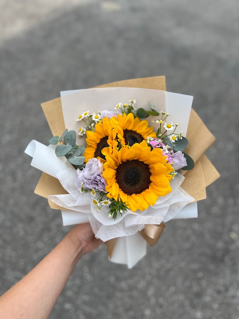 A hand holds a vibrant bouquet of sunflowers, wrapped in brown and white paper.