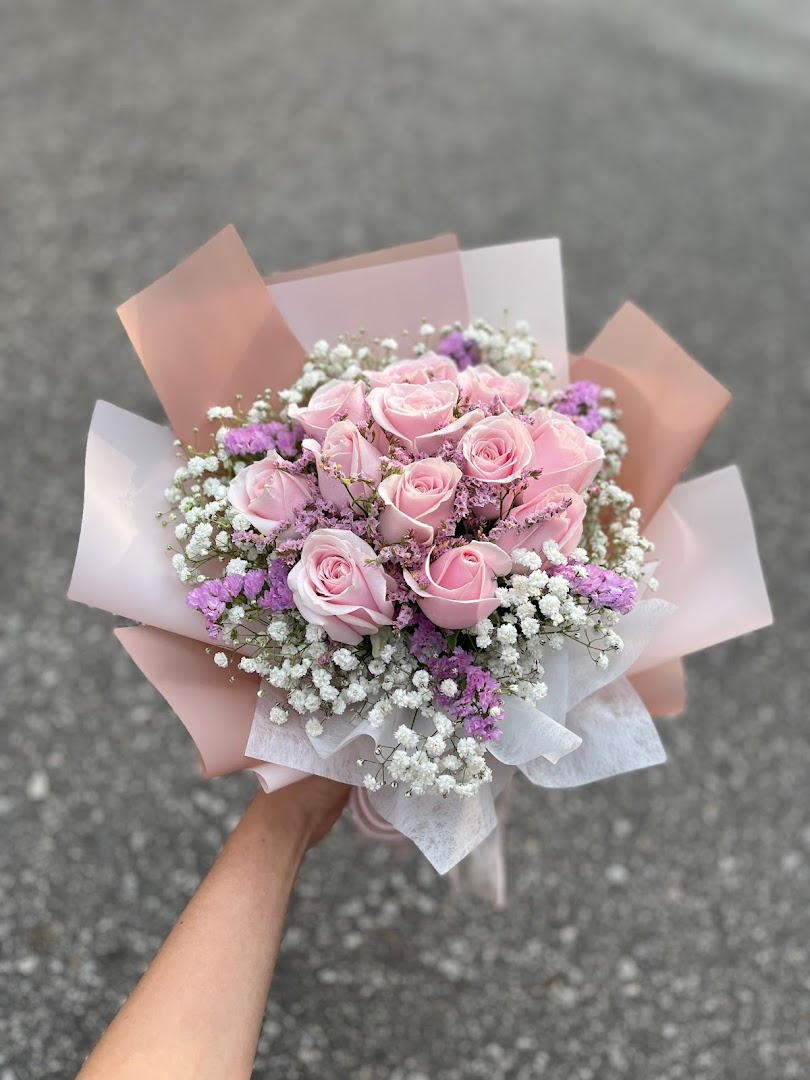 A hand holds a beautiful bouquet of pink roses, baby's breath, and purple filler flowers, wrapped in elegant paper.