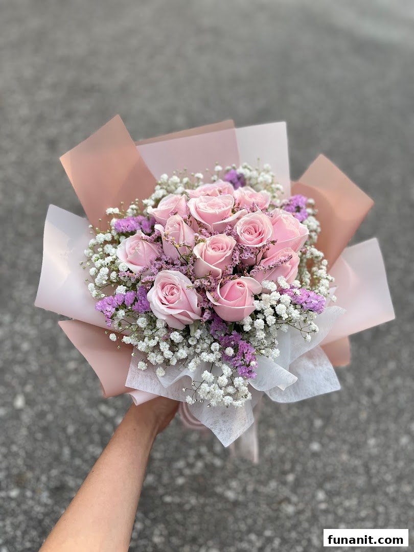 A hand holds a beautiful bouquet of pink roses, baby's breath, and purple filler flowers, wrapped in elegant paper.