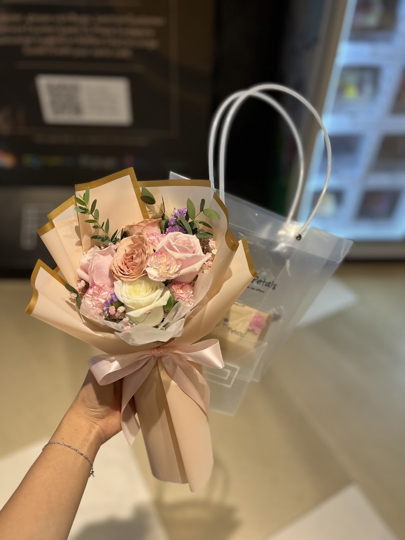 A hand holds a beautiful bouquet of pink and white flowers, wrapped in peach paper, with a clear bag in the background.