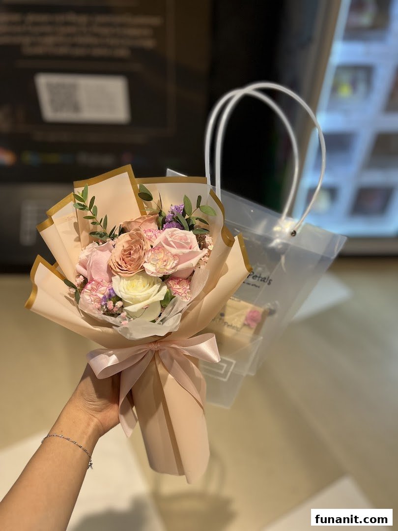 A hand holds a beautiful bouquet of pink and white flowers, wrapped in peach paper, with a clear bag in the background.